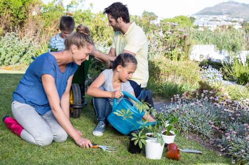 A family is gardening in a well-maintained outdoor space with a lush green lawn surrounded by flower beds and shrubs. The foreground features two children, one girl in a blue backpack and a woman, crouched down and planting a young shrub with gardening tools and pots nearby. Behind them, a man is standing, observing and smiling, while another boy in a striped shirt kneels in the background. The garden includes a variety of plants, with some flowering, and a paved pathway can be seen in the distance. The scene is set on a bright, sunny day, with clear skies and natural light illuminating the vibrant greenery and floral colours. This outdoor area, likely part of a residential garden in Hackbridge, demonstrates typical landscaping with grass, flower borders, and gardening activity, suitable for outdoor maintenance and gardening services offered by Gardening Services Hackbridge.