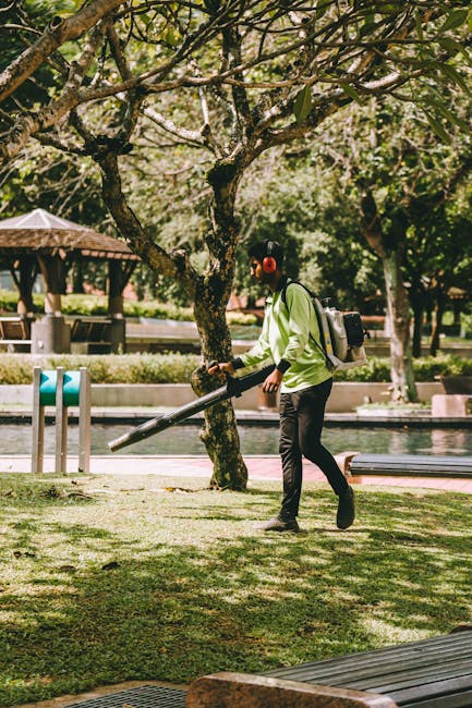 A person wearing headphones, a light green jacket, and black pants is using a leaf blower to clear fallen leaves from a well-maintained grassy area in a park. They are carrying a backpack-style blower