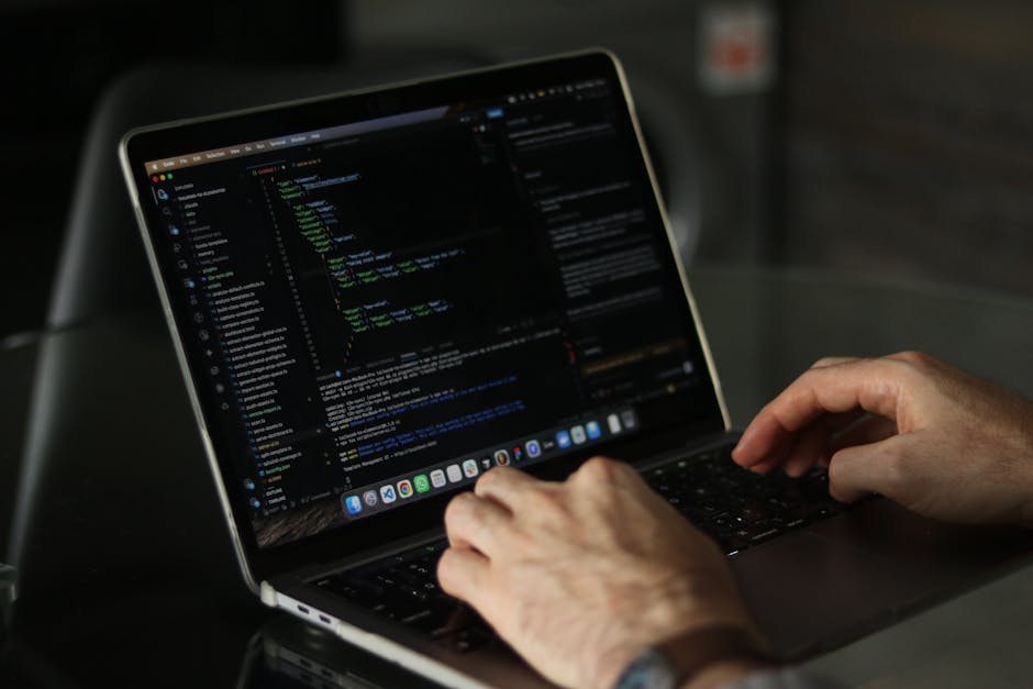 A close-up view of a person's hands typing on a laptop keyboard, with the screen displaying a dark code editor interface filled with lines of programming code in various colors. The laptop is placed o